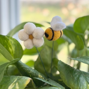 May include: A felt flower and bee decoration. The flower has white petals and a yellow center. The bee is yellow with black stripes and white wings. The flower and bee are attached to green wire stems, set against a backdrop of green leaves.