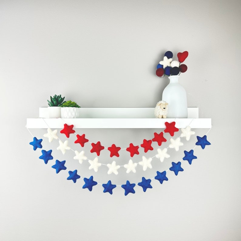 May include: A white shelf with a red, white, and blue felt star garland hanging from it. The garland is made of felt stars in three sizes and is strung on white twine. There is a small potted plant and a white vase with a small white sheep figurine on the shelf.