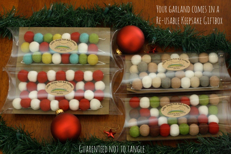 May include: Four clear plastic boxes with felt ball garlands inside. The garlands are red, white, green, and brown. The boxes are on a wooden surface with green garland around the edges. The text "Your garland comes in a re-usable keepsake giftbox" is at the top of the image. The text "Guaranteed not to tangle" is at the bottom of the image.