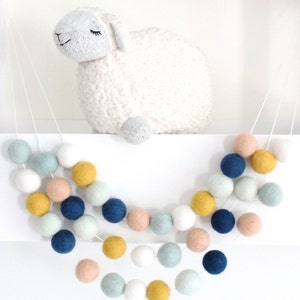 May include: A garland of felt balls in shades of blue, green, yellow, and pink. The garland is strung on a white cord and hangs from a shelf. A white stuffed sheep is sitting on the shelf behind the garland.