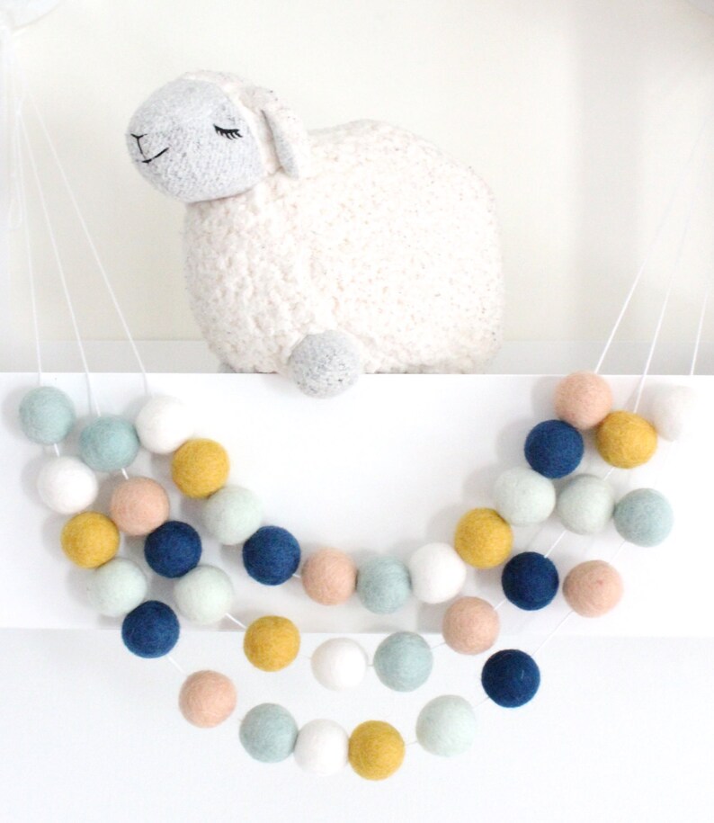 May include: A garland of felt balls in shades of blue, green, yellow, and pink. The garland is strung on a white cord and hangs from a shelf. A white stuffed sheep is sitting on the shelf behind the garland.