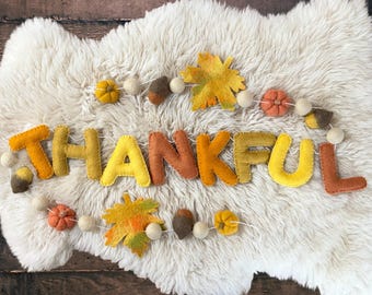 May include: A felt banner spelling out the word "THANKFUL" in shades of yellow, orange, and brown. The banner is decorated with felt leaves, pumpkins, acorns, and white pom-poms. The banner is displayed on a white, fluffy surface.