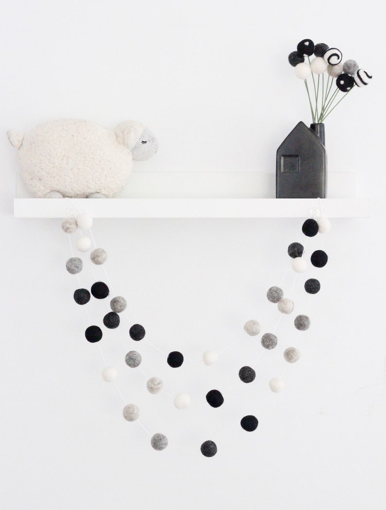 May include: A white shelf with a white sheep plush toy, a black house-shaped vase with black and white pom-poms, and a black and white pom-pom garland hanging from the shelf.