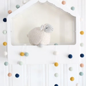 May include: A white shelf with a scalloped edge, decorated with a garland of colorful felt balls. A white stuffed sheep sits on the shelf. The garland hangs down from the shelf, with the felt balls in shades of blue, green, yellow, and pink.