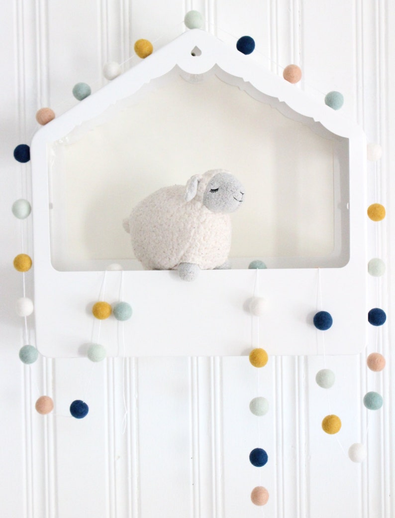 May include: A white shelf with a scalloped edge, decorated with a garland of colorful felt balls. A white stuffed sheep sits on the shelf. The garland hangs down from the shelf, with the felt balls in shades of blue, green, yellow, and pink.