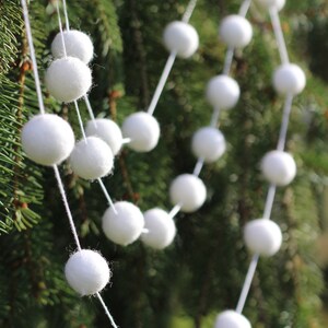 May include: A white felt ball garland hanging on a green evergreen tree branch. The garland is made of white felt balls strung together with white thread.