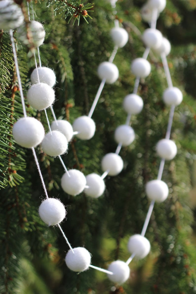 May include: A white felt ball garland hanging on a green evergreen tree branch. The garland is made of white felt balls strung together with white thread.