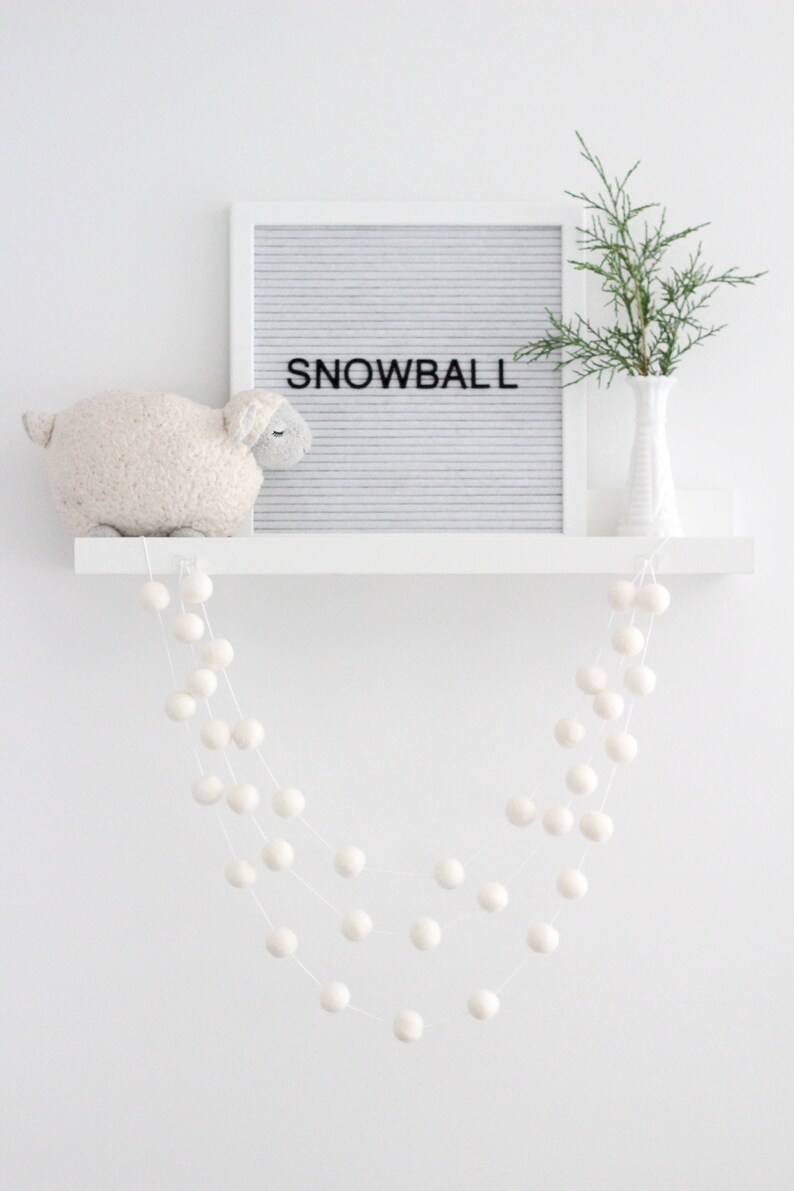 May include: White felt ball garland hanging from a white shelf with a white sheep plush toy and a letter board that says "SNOWBALL" on it.
