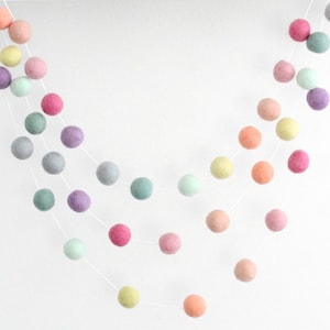 May include: A garland of pastel colored felt balls strung on white thread. The garland features shades of pink, purple, green, and yellow.