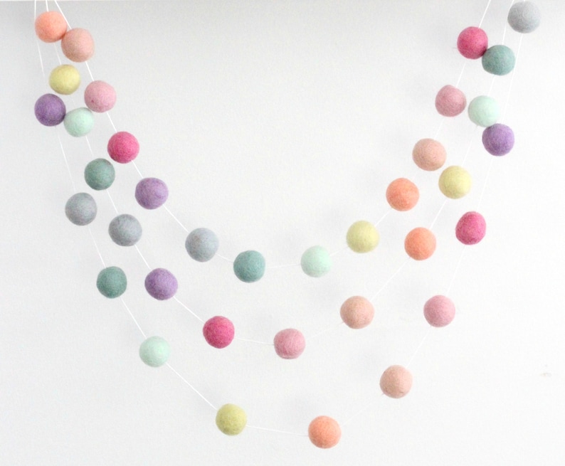 May include: A garland of pastel colored felt balls strung on white thread. The garland features shades of pink, purple, green, and yellow.