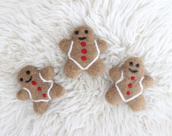 May include: Three gingerbread man ornaments made of felt. The gingerbread men are brown with white icing and red buttons. They are lying on a white fluffy surface.