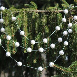 May include: A white pom-pom garland strung on a white string, hanging on a green evergreen tree branch.