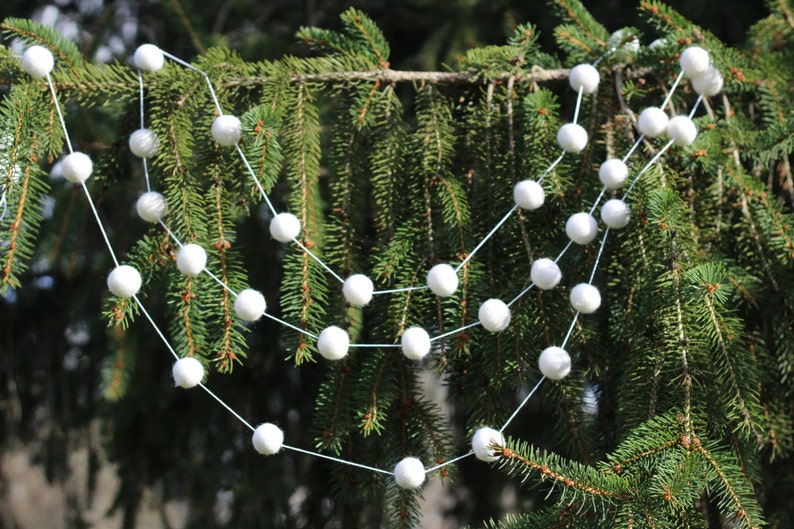 May include: A white pom-pom garland strung on a white string, hanging on a green evergreen tree branch.