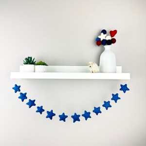 May include: A blue felt star garland hangs from a white shelf. The garland is made of 12 stars and is perfect for a nursery or child's room.