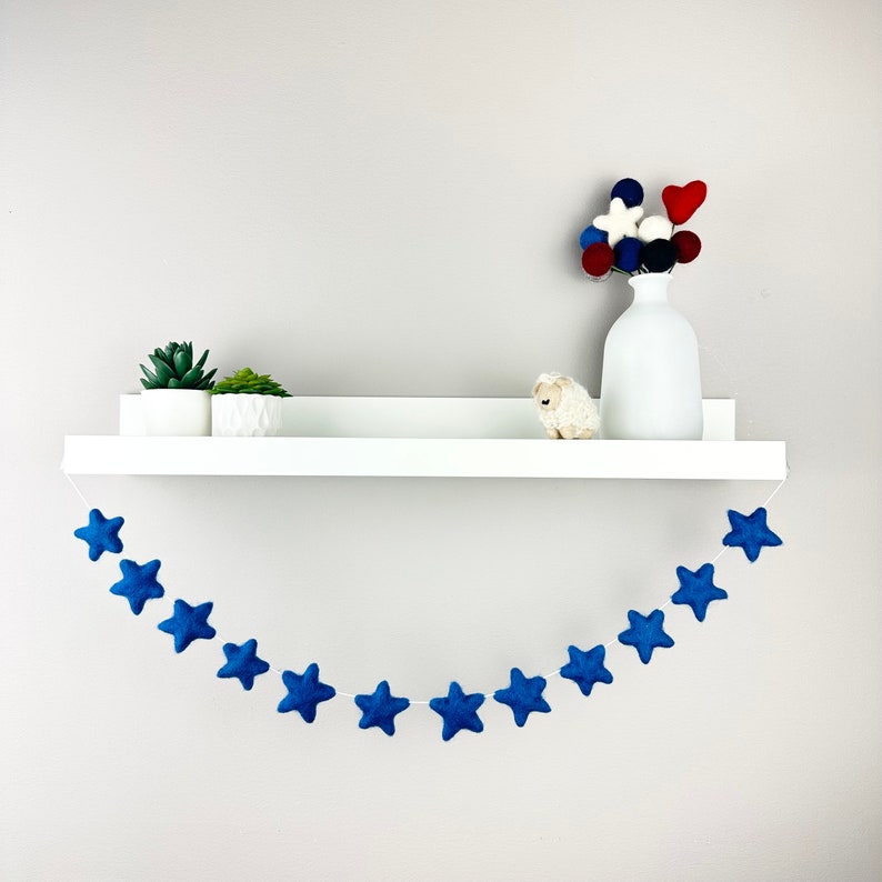 May include: A blue felt star garland hangs from a white shelf. The garland is made of 12 stars and is perfect for a nursery or child's room.