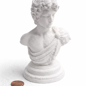 May include: A white bust of a classical figure, possibly a Greek or Roman god, with detailed facial features and draped clothing. The sculpture sits on a round base. A penny is next to the bust for scale.