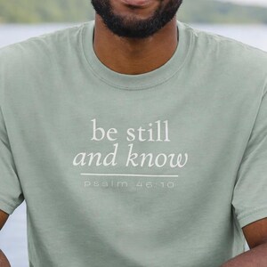 May include: A pale green t-shirt featuring the white text "be still and know". Beneath this, in smaller text, is "psalm 46:10". The shirt appears to be made of a soft fabric.