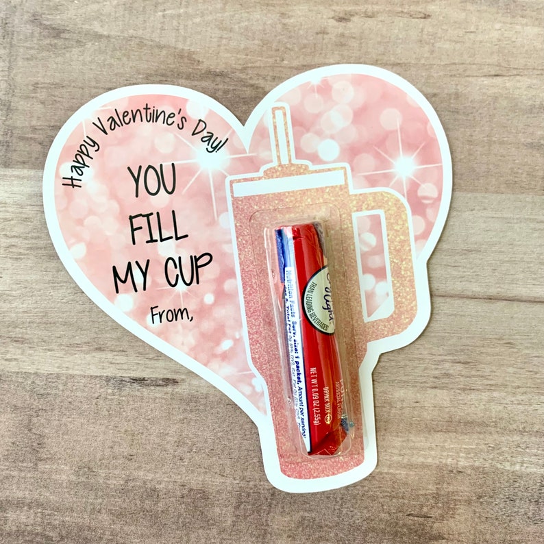 Puede incluir: Tarjeta de San Valent&iacute;n en forma de coraz&oacute;n rosa con purpurina y un vaso de pl&aacute;stico transparente con una pajita. La tarjeta dice "&iexcl;Feliz D&iacute;a de San Valent&iacute;n! T&uacute; llenas mi vaso" y tiene un espacio para un nombre. Un b&aacute;lsamo labial rojo y azul est&aacute; dentro del vaso.