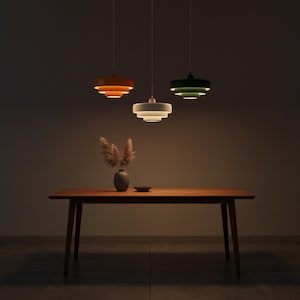 Peut inclure: Trois lampes suspendues à gradins, orange, blanche et verte, suspendues au-dessus d'une table en bois. Un vase avec des plantes séchées et un petit bol sont sur la table. L'ambiance de la pièce est chaleureuse et accueillante.