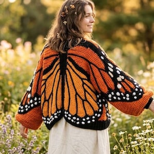 Könnte beinhalten: Ein handgefertigter Häkelpullover mit Schmetterlingsflügeln in Orange, Schwarz und Weiß. Der Pullover hat ein detailliertes Design, das die Flügel des Monarchfalters nachahmt. Die Person trägt den Pullover auf einer Blumenwiese.
