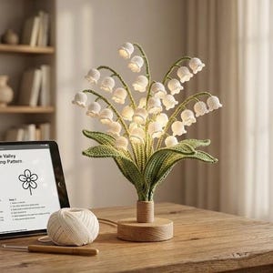 Könnte beinhalten: Eine gehäkelte Maiglöckchen-Lampe mit beleuchteten weißen Blütenköpfen und grünen Blättern. Die Lampe steht auf einem Holzsockel. Auf dem Tisch liegen ein Tablet mit einer Häkelanleitung, ein Knäuel Garn und eine Häkelnadel.