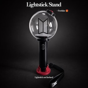 Peut inclure: Un support de bâton lumineux noir et rouge avec un dessus sphérique transparent contenant un logo noir. La base est rouge et le manche cylindrique est noir avec un petit logo. Le texte indique Lightstick Stand, + Freebies et Lightstick not Inclued.