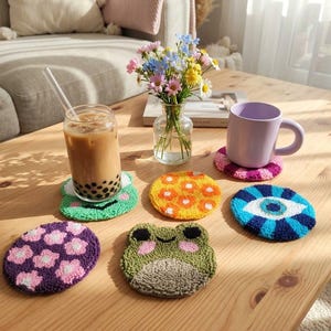 Pode incluir: Uma coleção de cinco porta-copos circulares tufados com vários designs, incluindo um sapo verde, um padrão floral amarelo, um olho azul, um padrão floral roxo e um segundo sapo verde, exibidos com um copo e uma caneca.