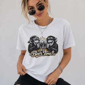 Op de afbeelding: Wit T-shirt met een cartoonontwerp van twee apen die proosten met bier, met de tekst "It's Beer Time!". De afbeelding is in zwart en geel. De persoon draagt een zonnebril en een zwarte jeans.