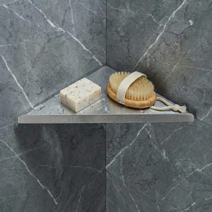 May include: A stainless steel corner shelf holds a bar of soap and a wooden brush with a white strap. The shelf is mounted on a gray marble wall, creating a spa-like atmosphere. The soap is rectangular and speckled.