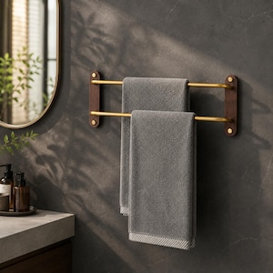 May include: A double towel rack featuring two horizontal brass-toned metal bars mounted between two dark brown wooden wall brackets, holding two folded gray towels with a textured border detail at the bottom edge of each towel.