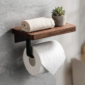 Può includere: Un moderno accessorio per il bagno con un ripiano in legno scuro e una staffa in metallo nero. Un rotolo di carta igienica bianca è appeso sotto. Un asciugamano beige arrotolato e una piccola pianta grassa in vaso sono sul ripiano.