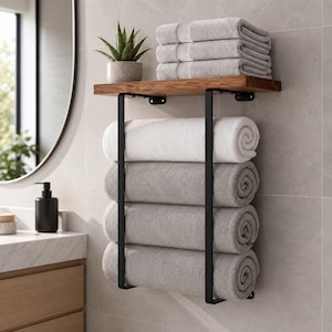 Può includere: Un mobiletto da bagno a parete con una mensola in legno naturale sostenuta da due staffe in metallo nero. La mensola ospita una pianta succulenta in vaso, mentre la struttura metallica sottostante contiene quattro asciugamani arrotolati.