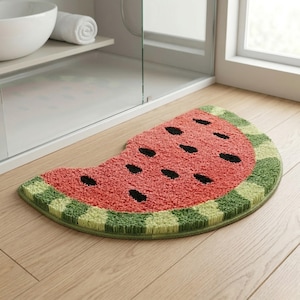 Puede incluir: Una alfombra de baño con forma de sandía. El centro es rojo con detalles de semillas negras, y el borde es verde con un diseño a cuadros. La alfombra está colocada sobre un suelo de madera, cerca de un mueble de baño y una ventana.