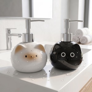 Pode incluir: Dois dispensadores de sabonete em forma de gato. Um é branco com orelhas castanhas e um pequeno nariz e olhos pretos. O outro é preto com olhos brancos. Ambos têm tampas de bomba prateadas e estão sobre uma superfície branca com gotas de água.