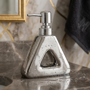 Pode incluir: Um dispensador de sabonete cromado com um design triangular único e uma bomba integrada. A superfície polida e reflexiva é adornada com gotas de água. Está colocado sobre uma bancada de mármore. O design inclui um vazio triangular.