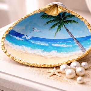 May include: A large seashell painted with a beach scene, featuring a palm tree, ocean waves, and sandy shore. The shell's edge is trimmed with gold paint. Pearls and a starfish are scattered nearby.