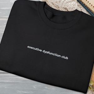 May include: Black crew neck sweatshirt with the text "executive dysfunction club" in white. The sweatshirt is displayed on a light gray wooden surface. The text is centered.