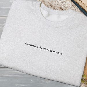 May include: A pale grey sweatshirt featuring the phrase "executive dysfunction club" in black lettering. The casual crew neck top is made from a soft fabric, suitable for everyday wear.