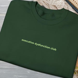 May include: Dark green crew neck t-shirt with the text "executive dysfunction club" in a light green font. The shirt is made of a soft material and is displayed on a wooden surface.