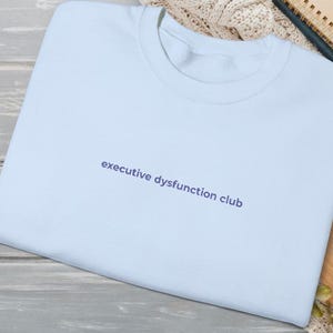 May include: A pale blue sweatshirt featuring the navy blue text "executive dysfunction club". The folded sweatshirt is on a wooden surface. The text is printed in a simple, sans-serif font and is centered.