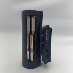 Può includere: Un portamunizioni cilindrico nero caratterizzato da un'apertura verticale che rivela quattro cartucce metalliche con punte color rame. Il supporto è fissato lateralmente a un meccanismo di clip da cintura in plastica nera per stoccaggio e accesso.