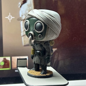 May include: A detailed figurine of a character in a gas mask and wrapped head covering. The figure is primarily gray and green, with a tan base. It is standing on a small platform.