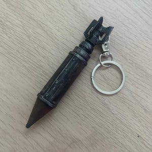 Könnte beinhalten: Ein dunkelbrauner und schwarzer Schlüsselanhänger in Form einer Bombe mit spitzer Spitze und gefiedertem Heck. Das Objekt hat einen zylindrischen Körper mit metallisch wirkenden Bändern und einen silberfarbenen Metallring mit Karabiner.