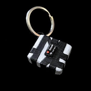 May include: A square-shaped keychain featuring a black and white striped pattern. The center of the object displays a small rectangular control panel with a single red button and two silver-colored accents, attached to a circular metal ring.