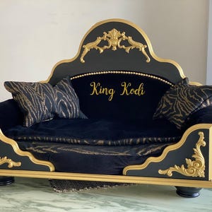 May include: A black and gold dog bed with a decorative headboard and ornate trim. The bed features a plush black cushion and two matching pillows with a gold and black zebra print. The words "King Kodi" are written in gold script.