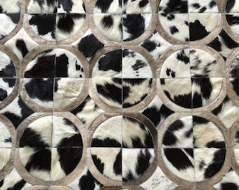 May include: A rug composed of circular patches of black and white cowhide leather. Each circular section is divided into four quadrants and framed by a light tan border, creating a repeating geometric pattern across the entire surface.