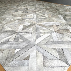May include: A white geometric patterned rug made of leather. The rug is made up of various sized squares and rectangles that are sewn together to create a geometric pattern.