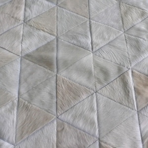 May include: A rug composed of numerous triangular patches of off-white and light gray animal hide. The pieces are stitched together in a geometric pattern, creating a textured surface with varying shades of cream and silver throughout the design.