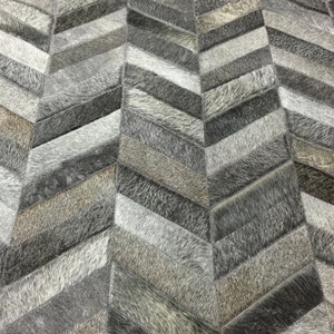 May include: A gray and white chevron patterned rug made from cowhide. The rug is made up of different sized pieces of cowhide sewn together to create a chevron pattern.