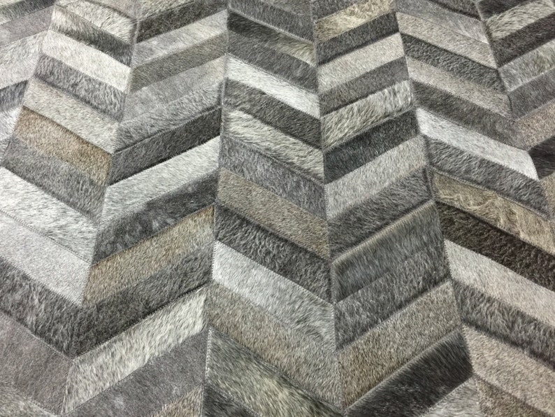 May include: A gray and white chevron patterned rug made from cowhide. The rug is made up of different sized pieces of cowhide sewn together to create a chevron pattern.